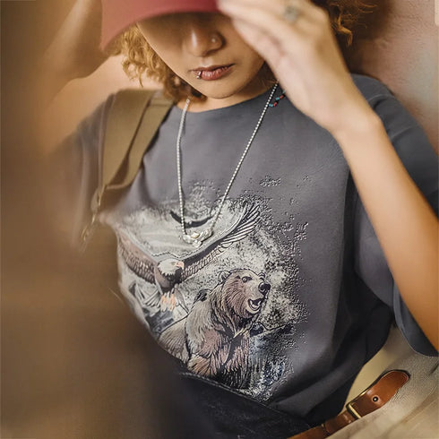 Person wearing a t-shirt with a bear and eagle design, holding a red hat.
